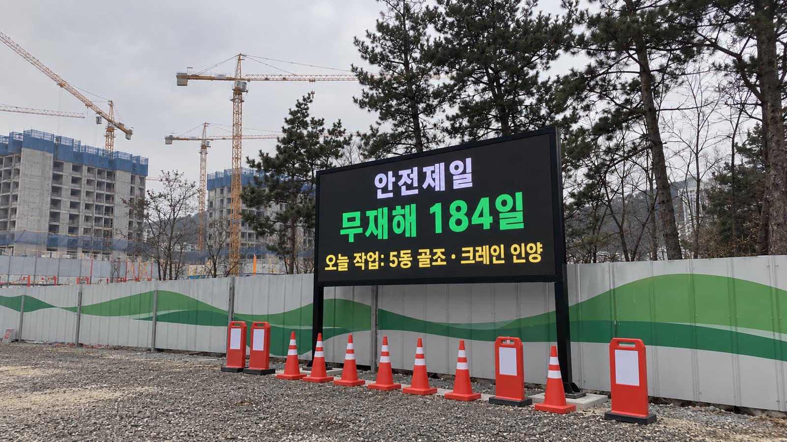 아파트 신축 공사현장 가설 울타리 앞 두 기둥 지주형 LED 안전전광판에 안전제일 무재해 일수와 오늘 작업이 표시된 장면