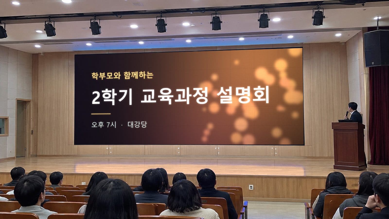 강당 무대 LED 전광판에 학부모와 함께하는 2학기 교육과정 설명회 시작 화면이 떠 있고 연단의 발표자와 객석이 보이는 장면