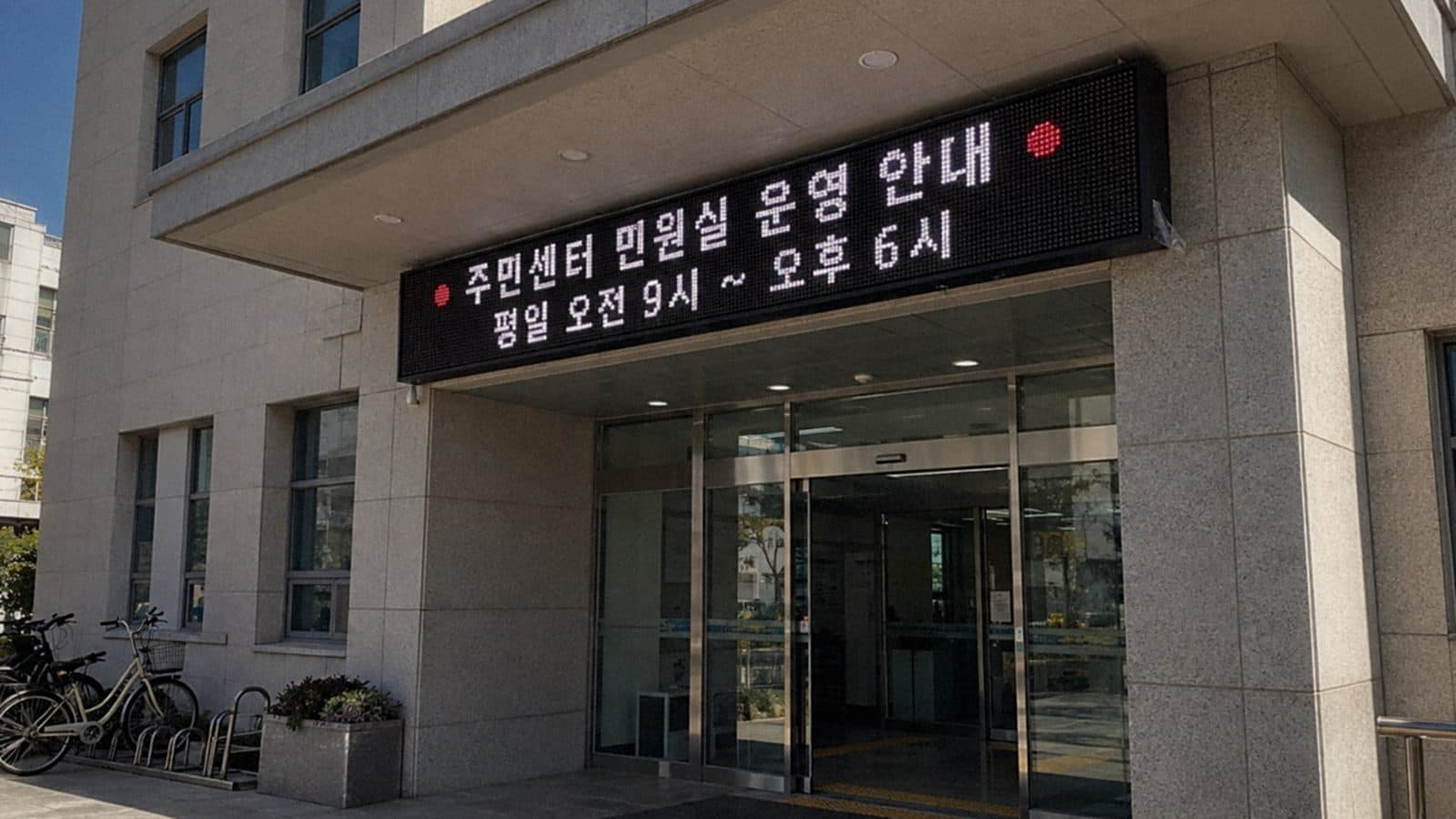 주민센터 현관 캐노피 위 외벽에 단 LED 전광판에 민원실 운영 시간 안내가 나오는 맑은 낮 장면