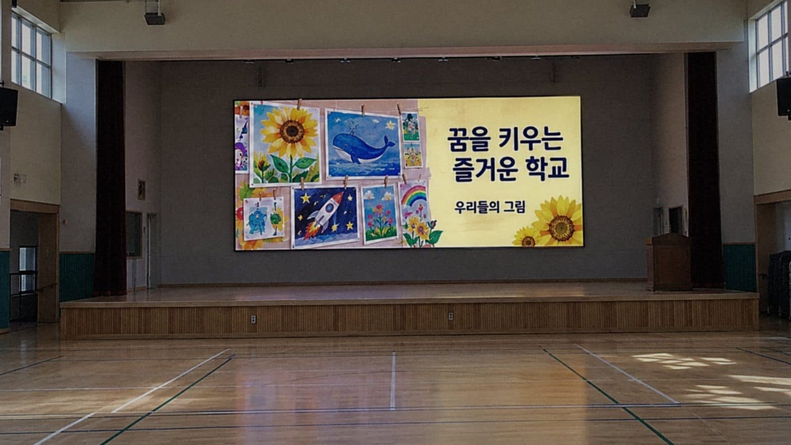 초등학교 강당 무대 뒷벽에 평평하게 붙인 실내 LED 전광판에 학생 그림과 학교 문구가 나오는 밝은 낮 장면