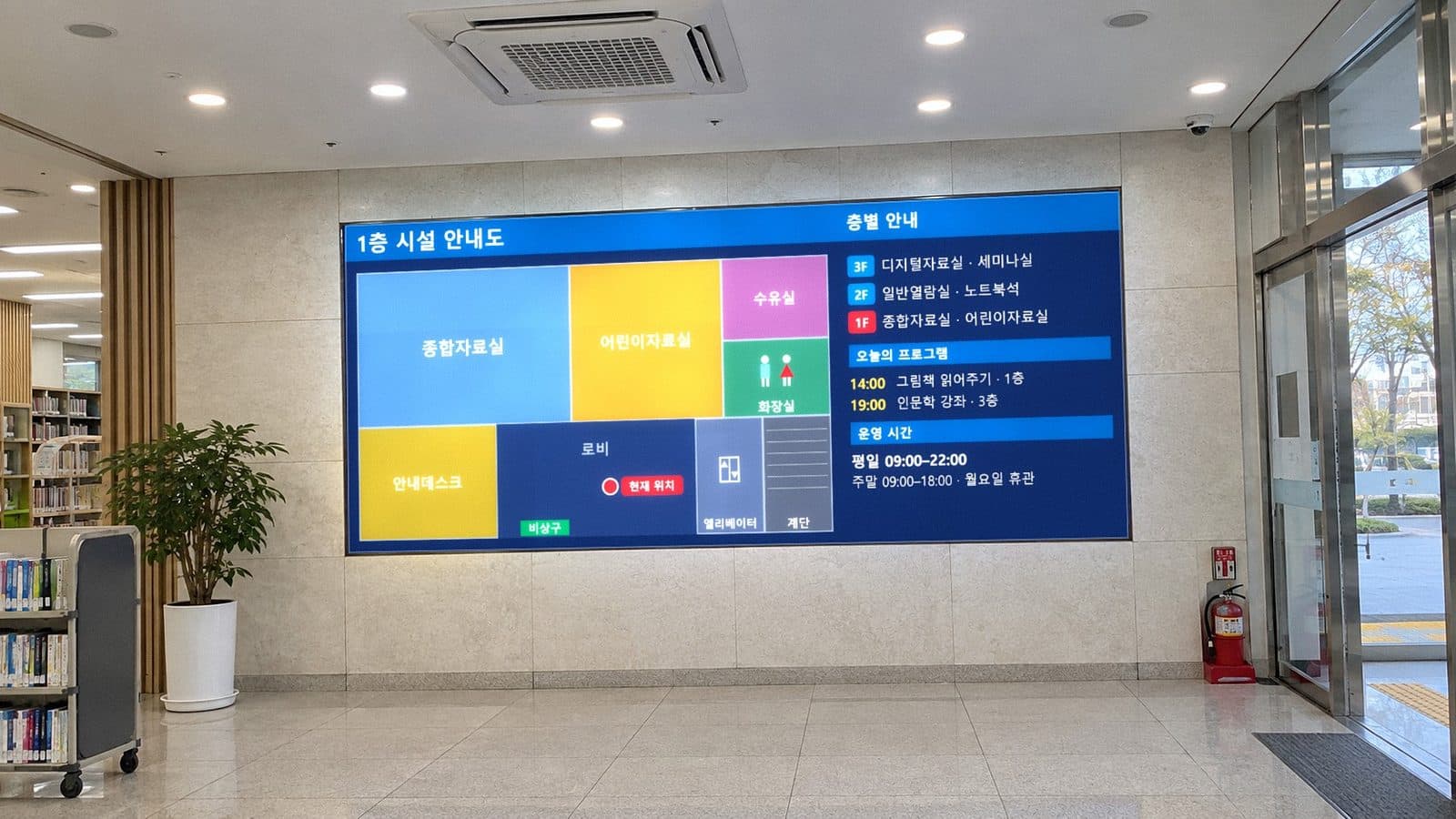 도서관 로비 벽면 LED 전광판에 1층 평면 안내도와 현재 위치, 층별 안내·오늘의 프로그램·운영 시간이 표시된 장면