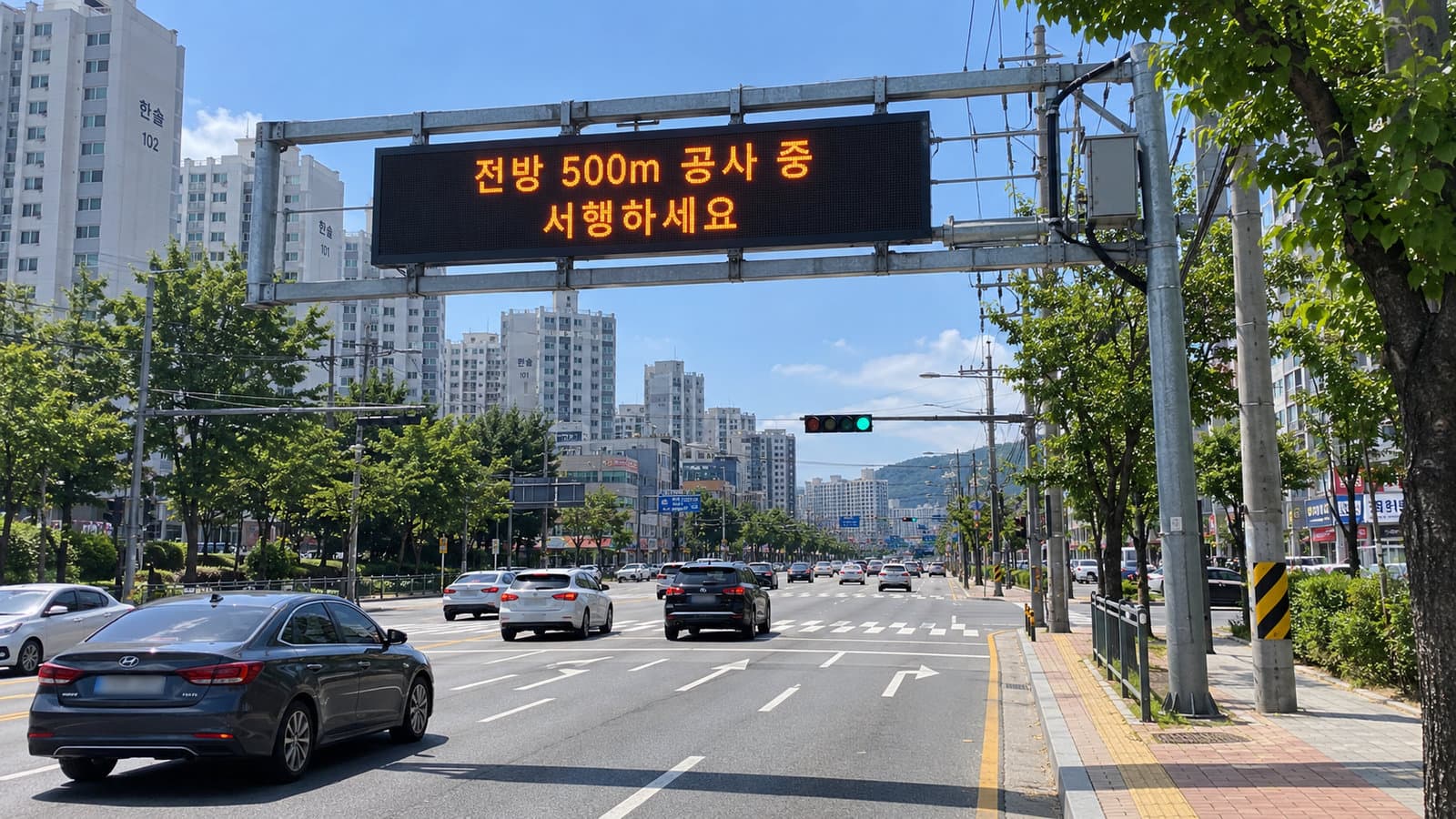 교차로 앞 도로 위 문형 지주에 설치된 도로전광표지에 전방 500m 공사 중 서행하세요 안내가 표시된 장면