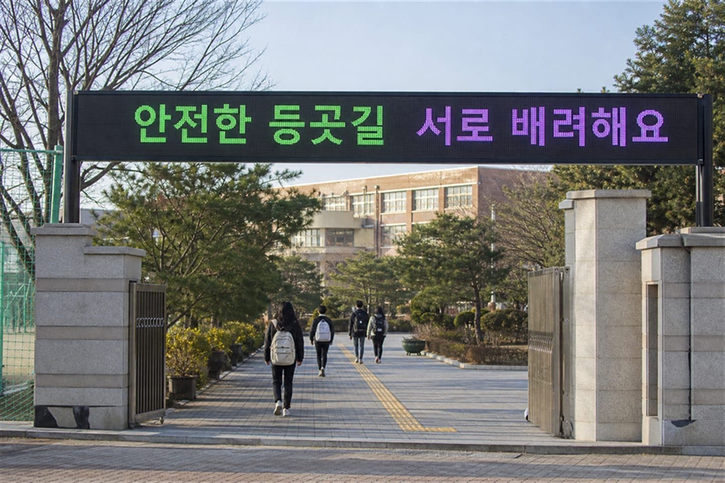학교 정문 아치에 설치된 가로형 전광판이 등굣길 안전 문구를 표시하고 있고 학생들이 걸어 들어가고 있다