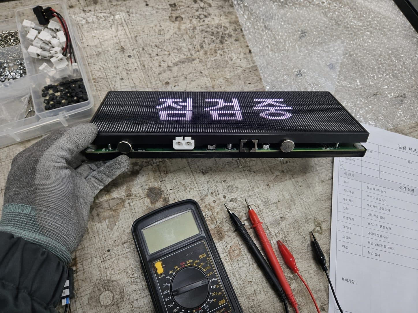 작업대 위에서 점검중 문구가 뜬 LED 모듈을 장갑 낀 손으로 들고 점검 체크시트와 대조하는 장면
