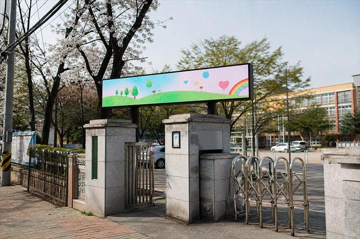 벚꽃이 핀 초등학교 정문 문주 위에 가로형 옥외 LED 화면이 설치되어 있다