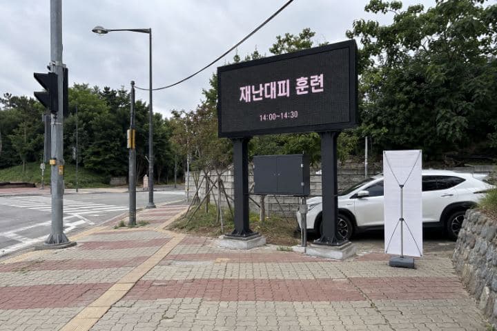 한국 교차로 인도 옆에 세워진 지주형 옥외 LED 표지가 재난대피 훈련 안내를 표시하고 있다