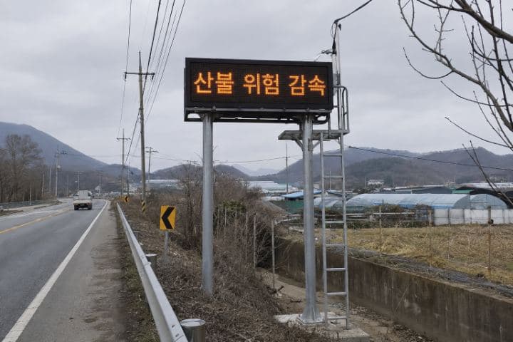 흐린 날 지방 도로변에 지주로 세워진 옥외 LED 화면에 "산불 위험 감속"이 표시되어 있다