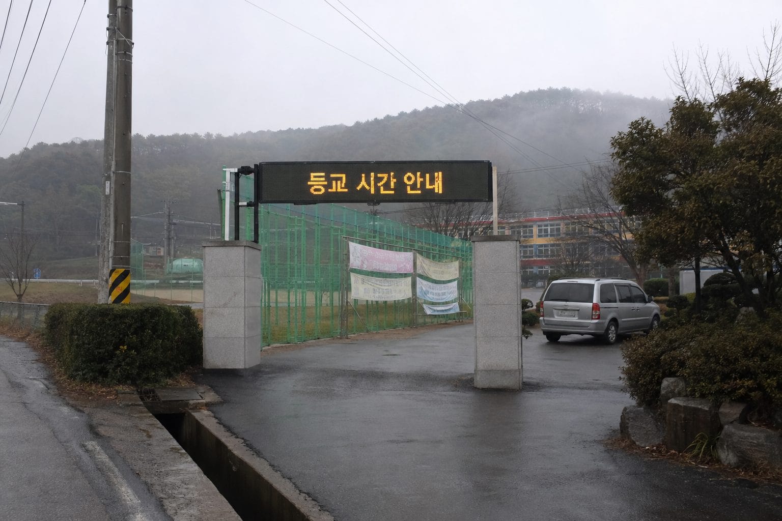 안개 낀 시골 학교 정문 위 가로형 전광판에 등교 시간 안내
