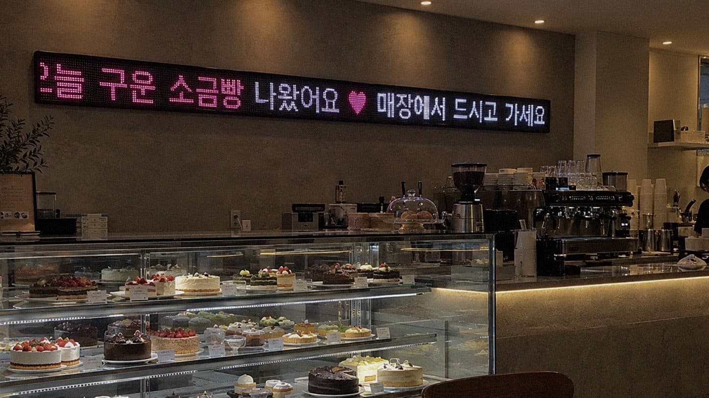 카페 케이크 쇼케이스 위 뒷벽에 단 띠형 LED 전광판에 오늘 구운 소금빵 나왔어요 문구가 나오는 장면
