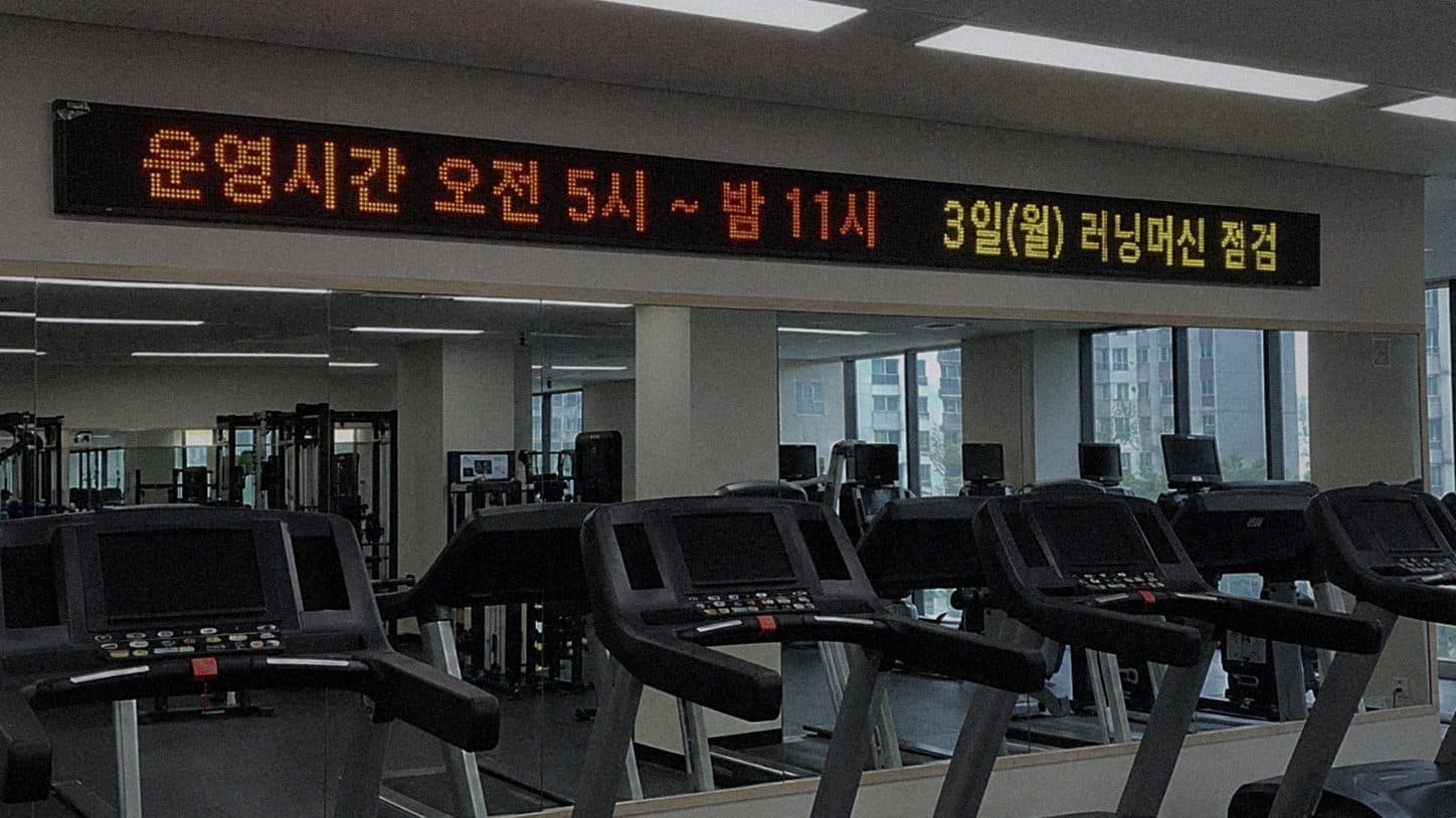 아파트 헬스장 거울 위 벽에 단 띠형 LED 전광판에 운영시간과 러닝머신 점검 안내가 나오는 장면